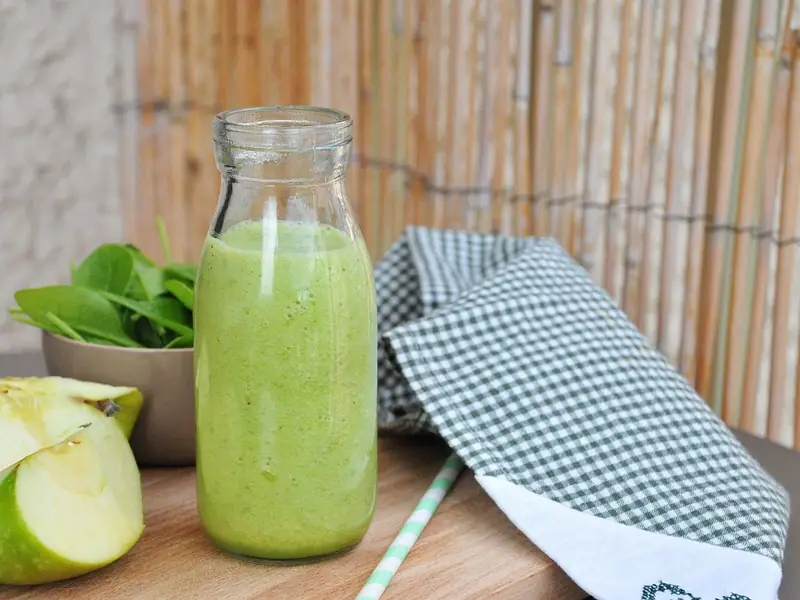 Πράσινο smoothie σε γυάλινο μπουκάλι γάλακτος πάνω σε ξύλινη επιφάνεια, με κομμένο πράσινο μήλο, ένα μπολ με σπανάκι, ένα ριγέ καλαμάκι και καρό πετσέτα.