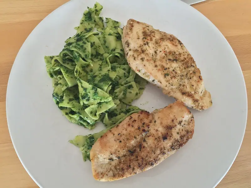 Zwei gewürzte, in der Pfanne gebratene Hähnchenbrustfilets neben einem Berg Bandnudeln in grünem Kräuterpesto auf einem weißen Teller auf einem Holztisch.