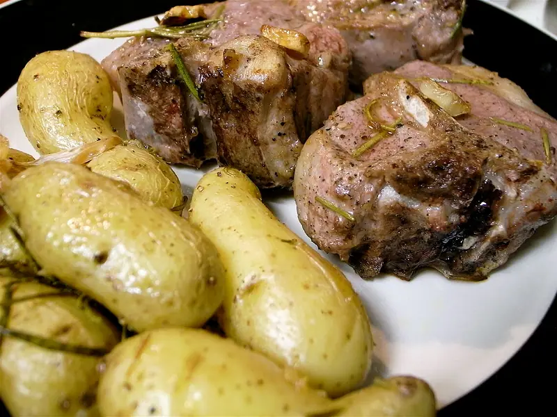 Seared lamb chops topped with rosemary sprigs and garlic slices on a white plate beside roasted fingerling potatoes.