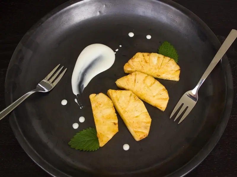 Golden pan-seared pineapple wedges on a dark plate with a swoosh of white cream, dots of cream, mint leaves and two forks