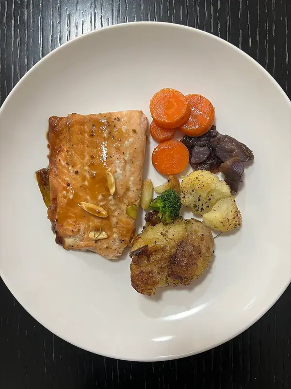Glazed salmon fillet with garlic slices on a white plate beside carrot rounds, a smashed potato, cauliflower, broccoli and purple potato.