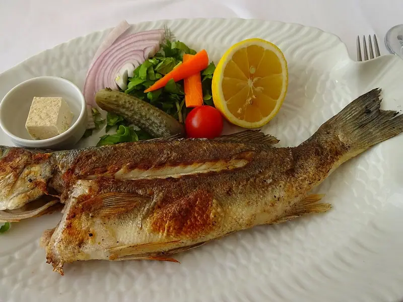 Bar entier grillé sur un plat blanc en forme de poisson, avec un demi-citron, de l'oignon rouge, de la verdure, de la carotte, un cornichon, une tomate cerise et du fromage frais à pâte blanche