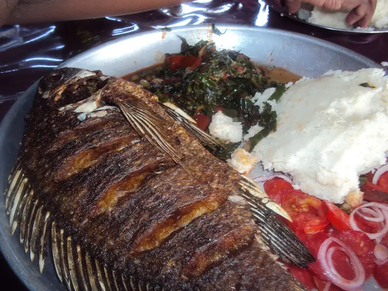 Whole charred tilapia on a metal platter with ugali, sautéed greens with tomato, and sliced tomatoes topped with red onion rings.