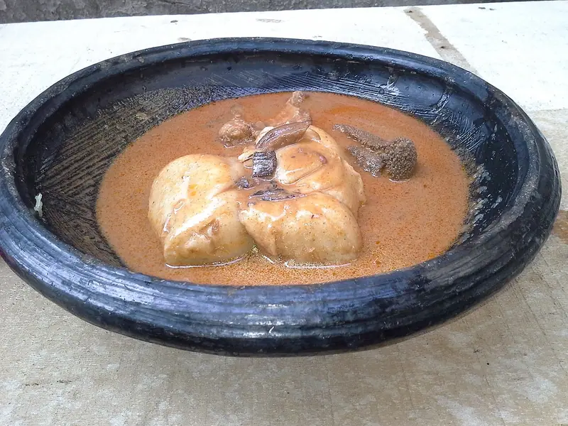 Orange jordnøddesuppe med lyse boller som fufu eller risboller og stykker af kød i en mørk, rustik lerskål på en stenoverflade.