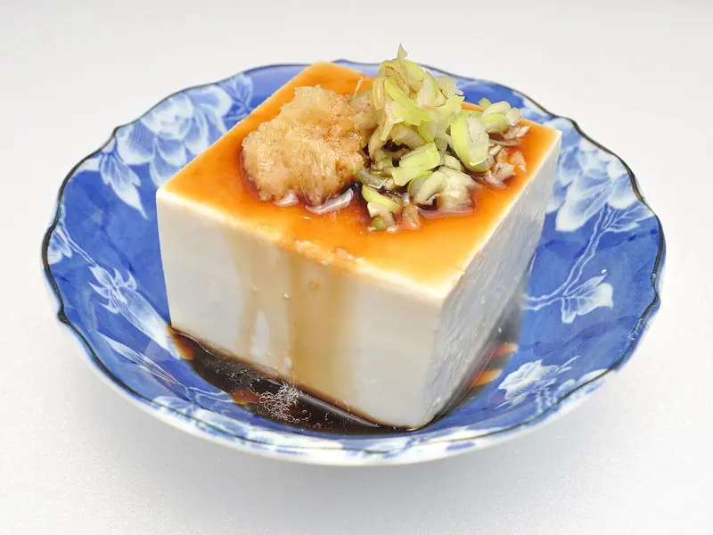 Un calup de tofu mătăsos alb, cu ghimbir ras și ceapă verde tăiată felii, stropit cu sos de soia, într-un bol albastru cu motive florale.