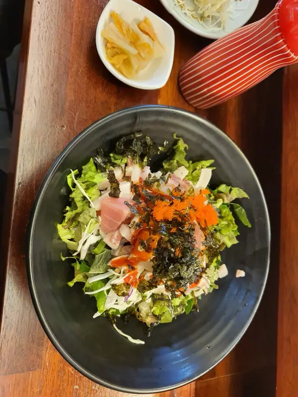 Black bowl of shredded lettuce and cabbage topped with raw tuna cubes, orange fish roe, seaweed flakes and red sauce, with side dishes
