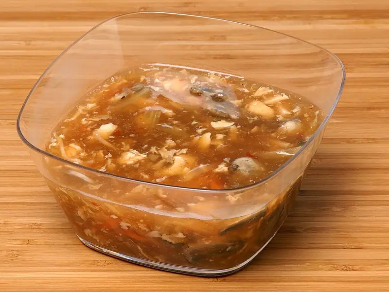 Thick brown hot and sour soup with tofu, egg ribbons, mushrooms and carrot strips in a clear square bowl on a bamboo board.