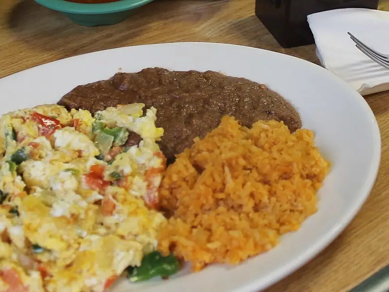 Ovos mexidos com tomate, cebola e pimenta verde em um prato branco ao lado de feijão refrito e arroz mexicano alaranjado sobre uma mesa de madeira.