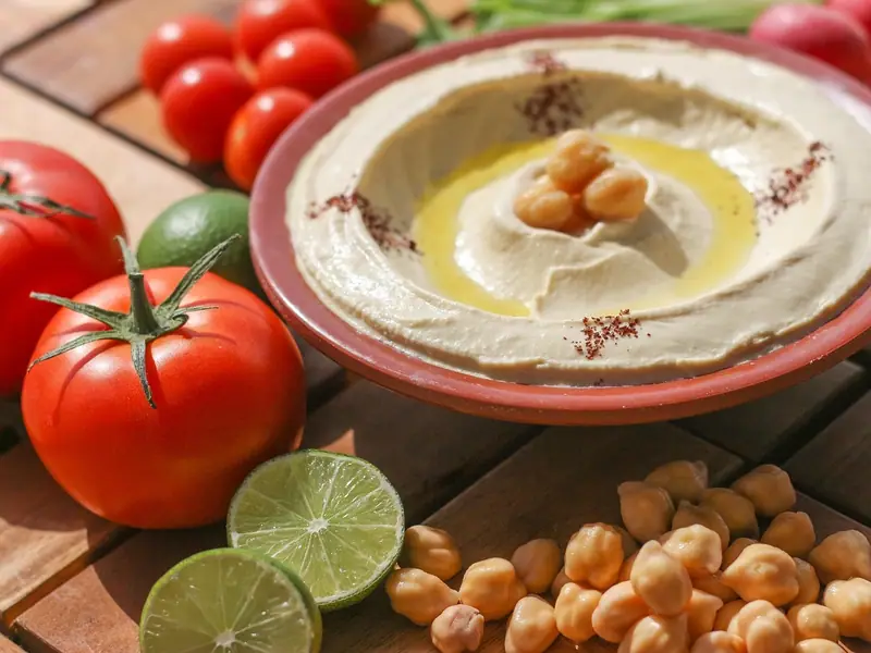 Krémový hummus v terakotové misce s olivovým olejem, celými cizrnami a sumakem, obklopený rajčaty, limetkami, ředkvičkami a cizrnou.