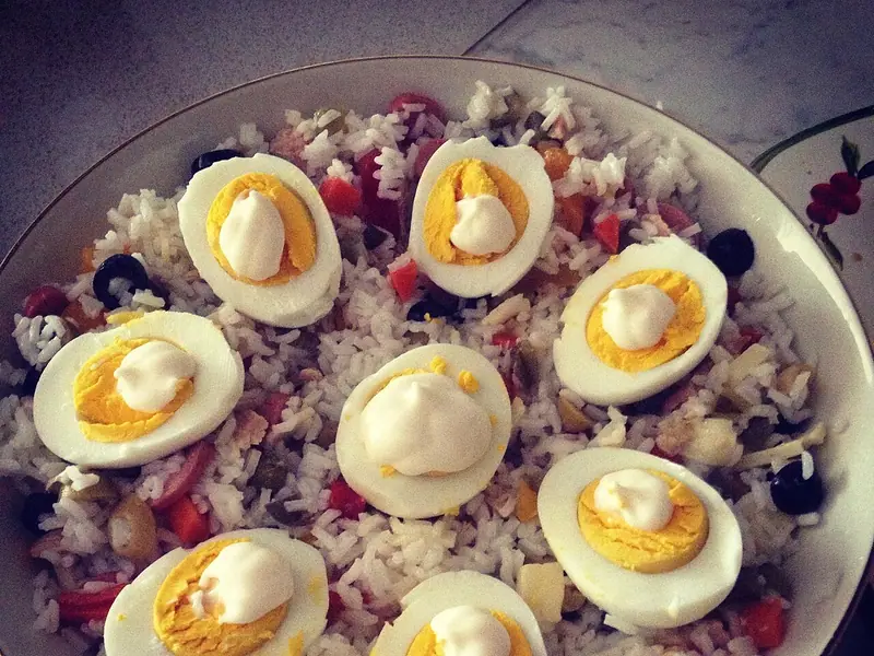 White rice salad with tuna, olives, red pepper and cheese cubes in a bowl, topped with eight halved boiled eggs with mayonnaise dollops