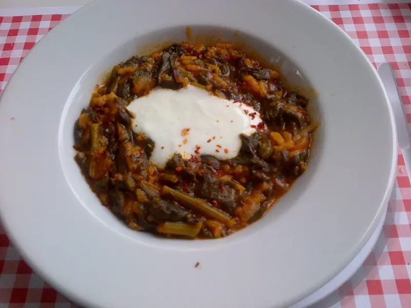 Spinatgryde med ris i en bred hvid skål, toppet med en klat hvid yoghurt og chiliflager, på en rødternet dug.