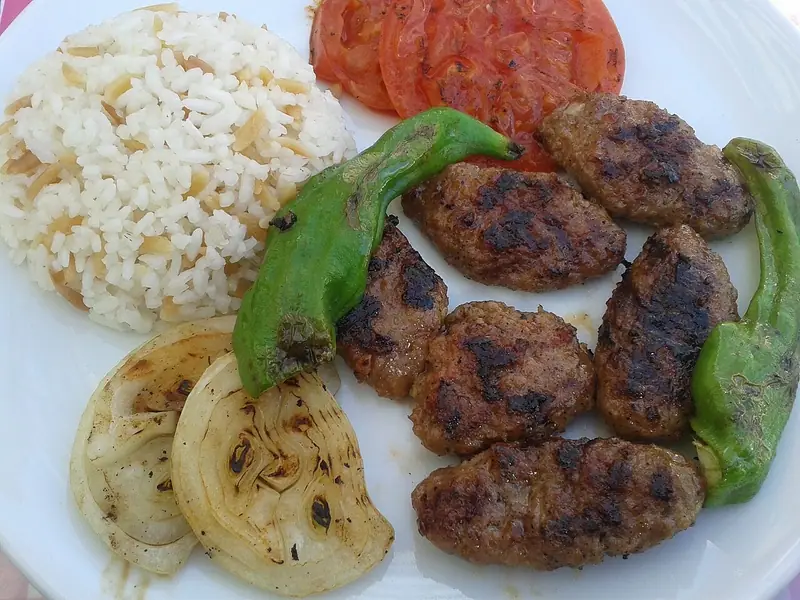 Charred oval köfte on a white plate with rice pilaf, grilled tomato slices, grilled onion wedges and blistered long green peppers.