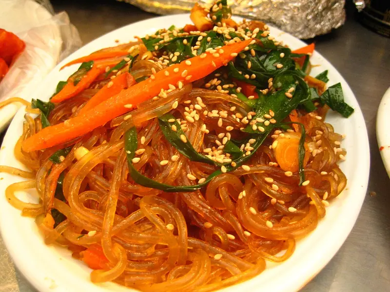 Glossy glass noodles on a white plate topped with carrot strips, wilted greens and a generous scattering of sesame seeds.