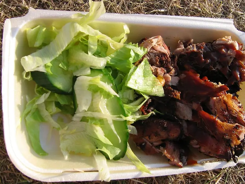 Charred jerk-style meat with a glossy brown sauce beside shredded lettuce and cucumber slices in a foam takeaway tray on dry grass.