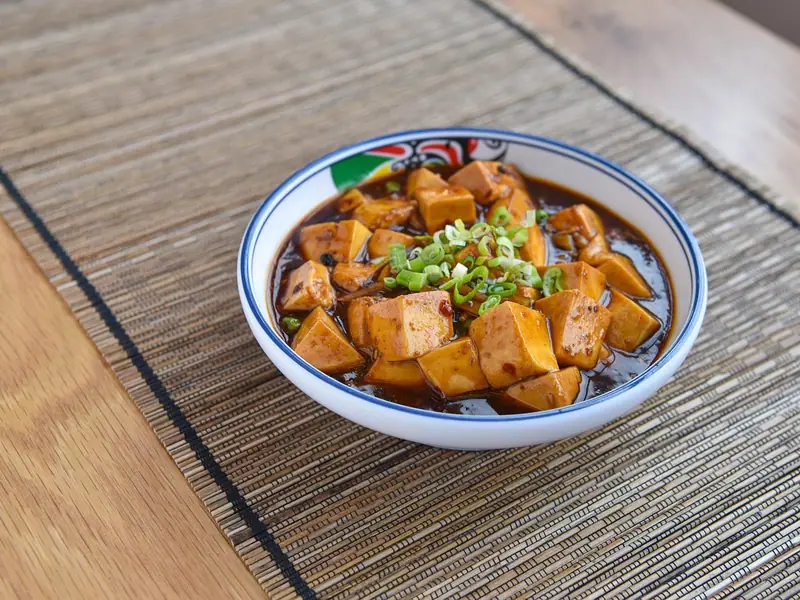 Cubes of tofu in a glossy red-brown chilli sauce topped with sliced scallions, served in a white bowl on a woven placemat.