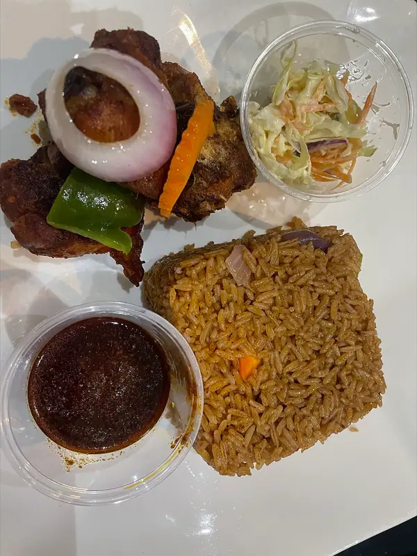 Orez Jollof pe o farfurie albă, cu pui prăjit, ornat cu un inel de ceapă, ardei verde și morcov, alături de boluri cu salată coleslaw și sos închis la culoare.