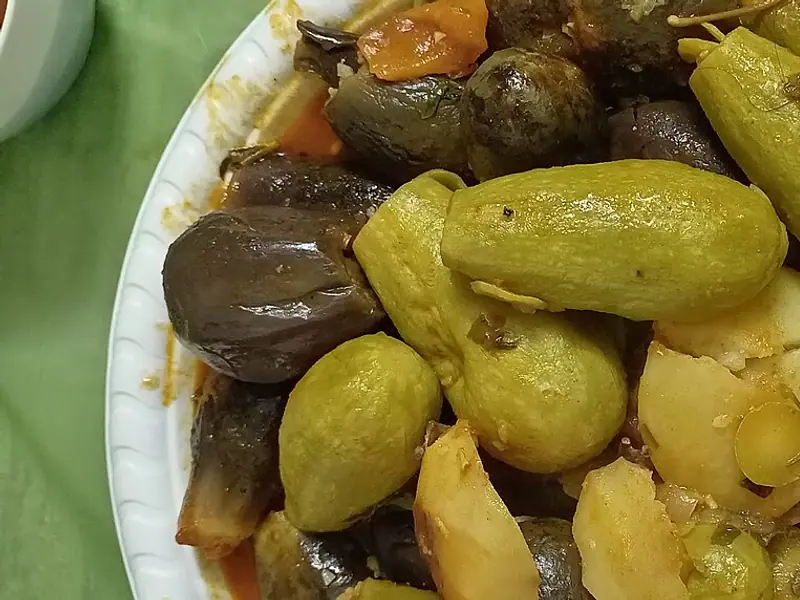 Yeşil bir örtünün üzerindeki beyaz karton tabakta, kırmızı domatesli suyun içinde açık yeşil kabak dolmaları, küçük koyu renkli patlıcanlar ve patates parçaları.