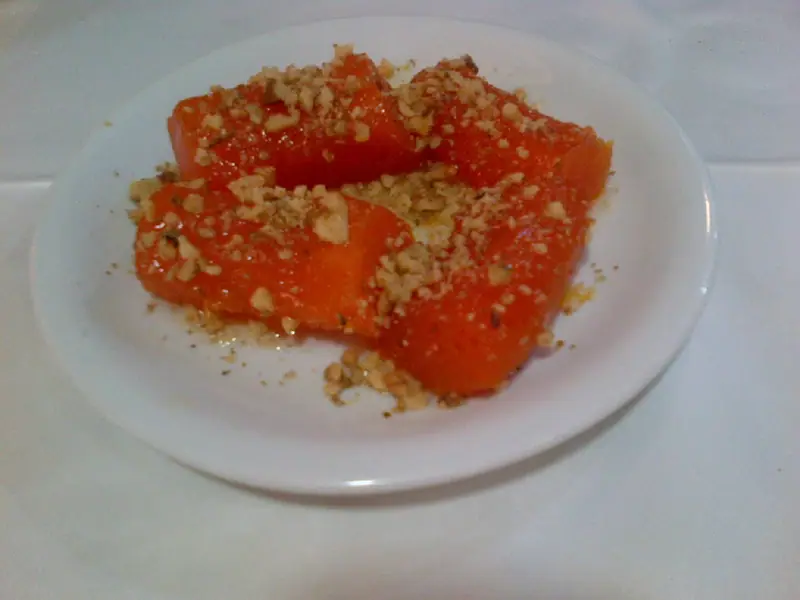 Glossy orange candied pumpkin pieces topped with finely chopped walnuts on a small white plate on a white tablecloth.