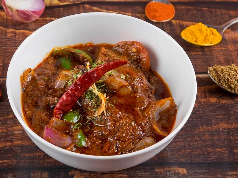 Hähnchencurry in einer dicken rotbraunen Masala mit grüner Paprika und roter Zwiebel, garniert mit einer getrockneten roten Chilischote, in einer weißen Schüssel auf Holz