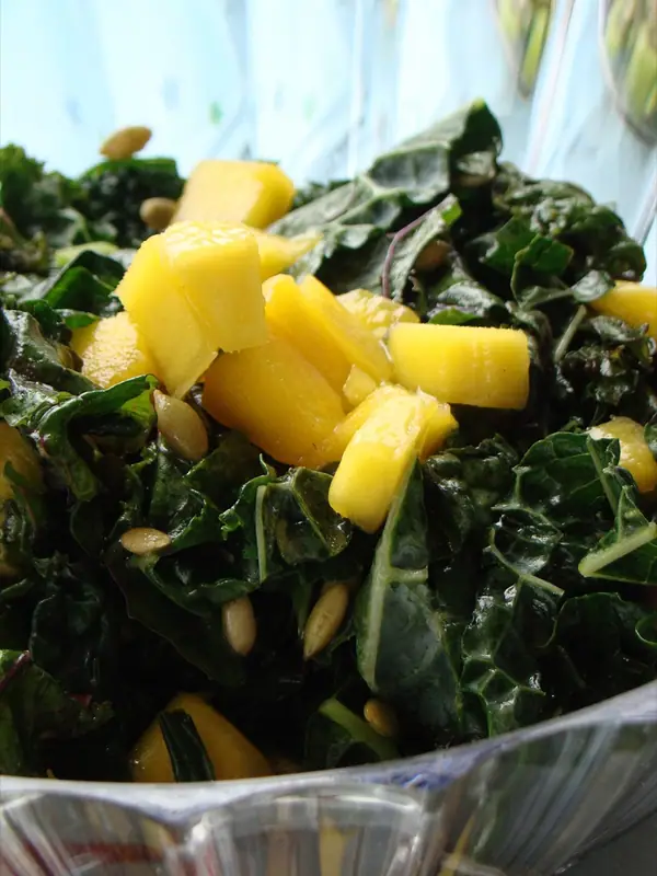 Ensalada de kale oscuro picado cubierta con dados de mango amarillo y semillas esparcidas, en un bol de cristal transparente.