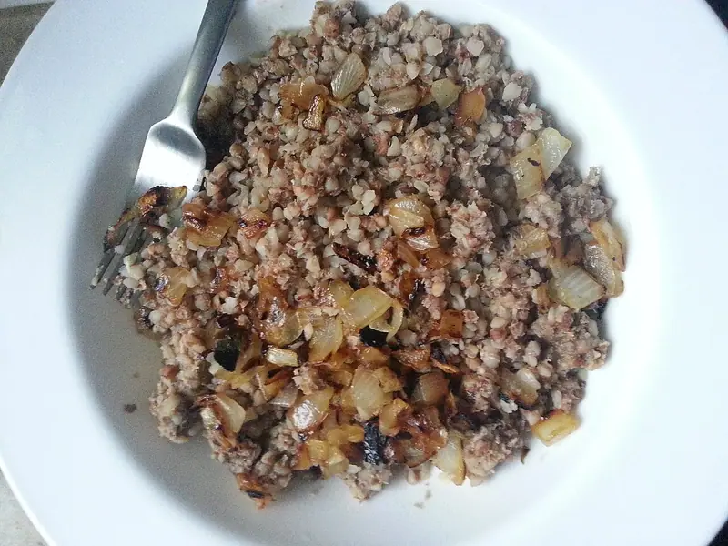 Cooked buckwheat kasha topped with golden fried onion pieces in a white bowl, with a fork resting on the left side