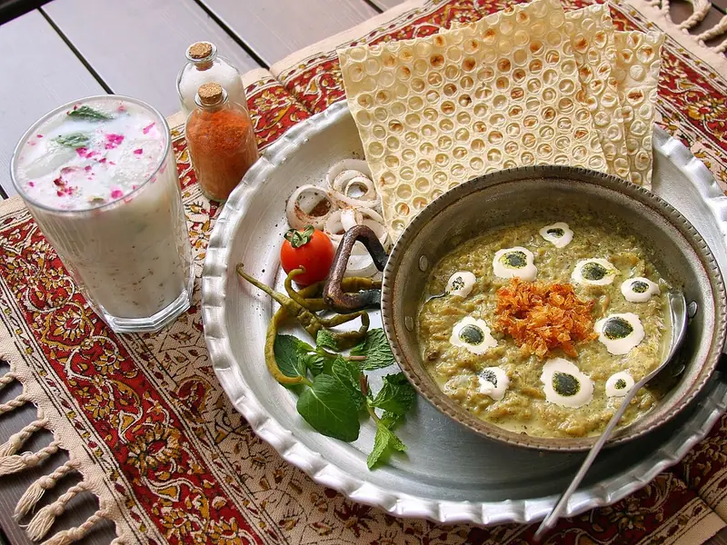 Bowl of creamy greenish stew topped with kashk dollops and fried onions, served with lavash, mint, chilies and a glass of doogh.