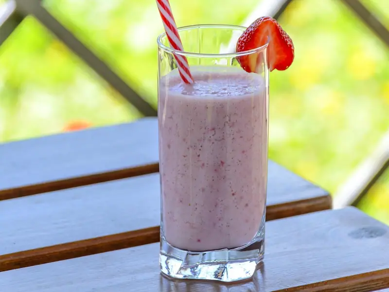 A tall glass of pink strawberry smoothie with a red-striped straw and a strawberry slice on the rim, on a wooden table outdoors.