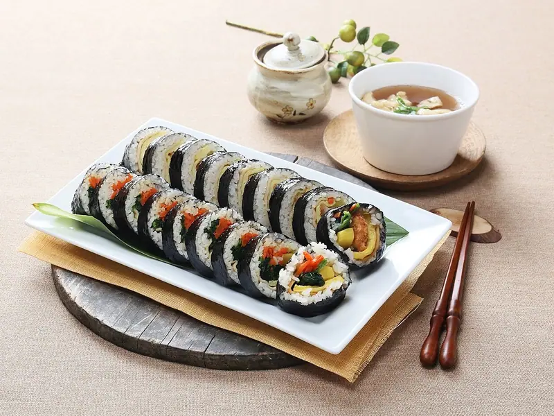 Sliced kimbap rolls with spinach, carrot, egg and yellow radish on a white platter, with a bowl of soup and wooden chopsticks nearby.