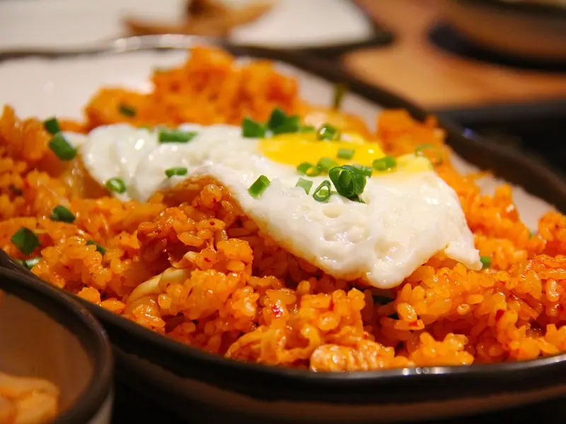 Orange kimchi fried rice on a dark plate, topped with a sunny-side-up fried egg and chopped green onions, with a side dish nearby.