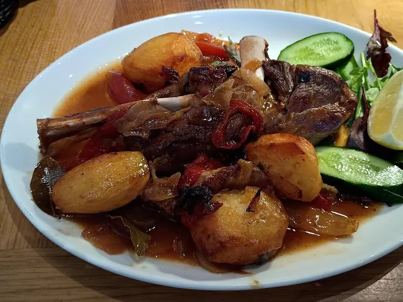 Braised lamb shank with golden roast potatoes, onions and red peppers in sauce, served with cucumber, salad leaves and a lemon wedge