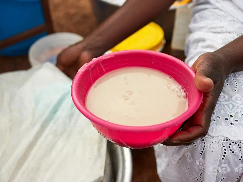 Hands holding a pink plastic bowl of thin, creamy beige porridge with small bubbles on top, outdoors beside a covered pot.