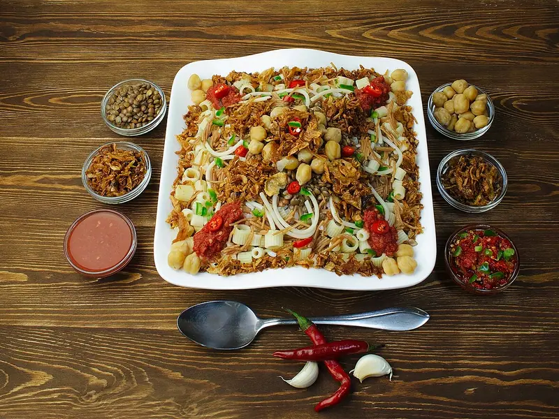 Koshari auf einer weißen quadratischen Platte, belegt mit Kichererbsen, gerösteten Zwiebeln, Tomatensauce und Chili, umgeben von kleinen Schälchen mit Toppings