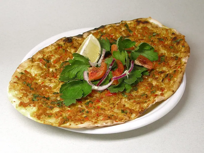 Thin oval lahmacun topped with spiced minced meat on a white plate, with parsley, red onion, tomato slices and a lemon wedge in the centre