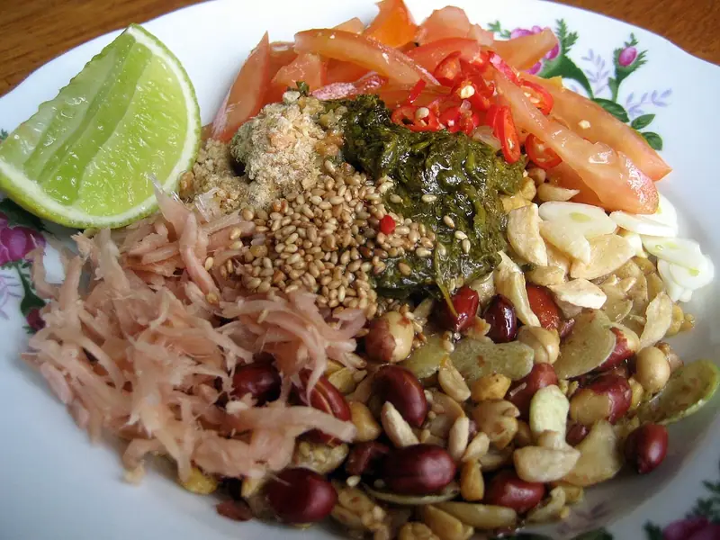 Tallerken med små bunker af fermenterede teblade, sesam, revet ingefær, tomat, rød chili, hvidløgschips, peanuts og friturestegte bønner med lime