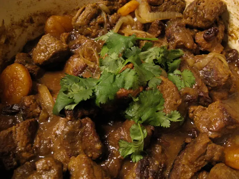 Nahaufnahme eines dunklen, saucigen Fleischeintopfs mit angebratenen Fleischstücken, Zwiebelscheiben, getrockneten Aprikosen und Rosinen, garniert mit frischen Korianderzweigen.