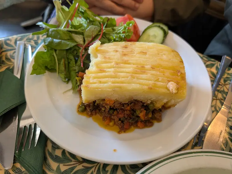 Et firkantet stykke shepherd's pie med gyldent kartoffelmos med gaffelriller over et fyld med ærter og gulerødder, serveret med grøn salat.
