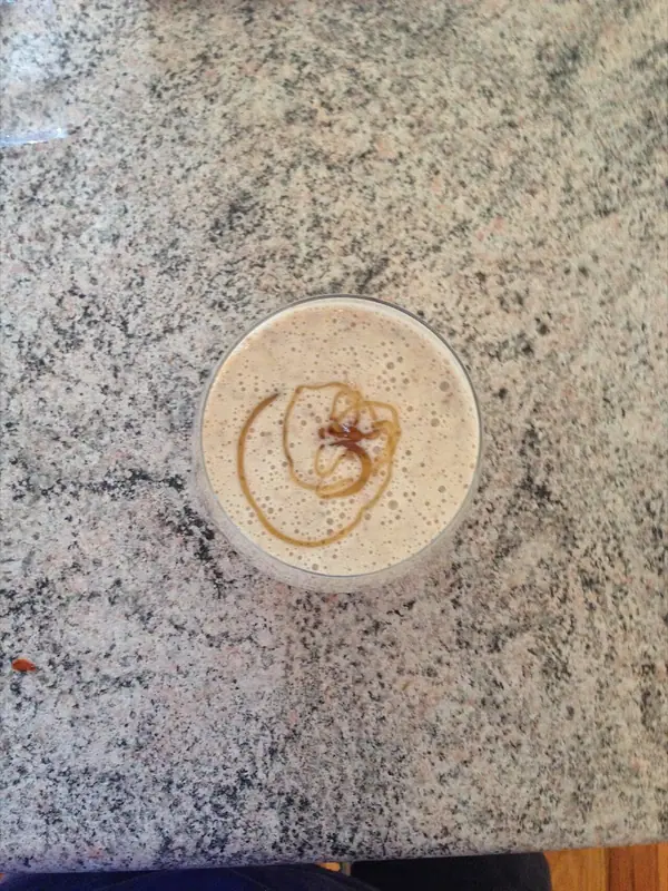 Draufsicht auf einen schaumigen, beigefarbenen Smoothie in einem klaren Glas mit einem karamellfarbenen Sirupwirbel obendrauf, auf einer Granitarbeitsplatte.