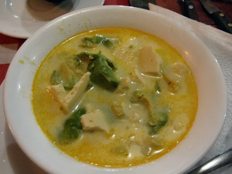 White bowl of thin, golden-yellow creamy broth with cubes of white cheese and chunks of avocado, set on a red tablecloth with napkin.