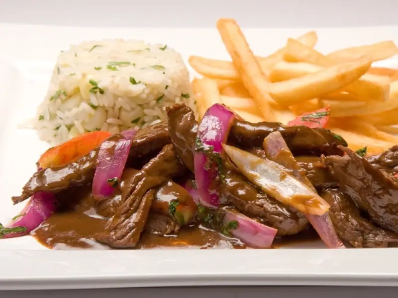 Strimler af oksekød i brun sauce med rødløg og tomat, serveret ved siden af en bunke hvide ris og gyldne pommes frites på en hvid tallerken.