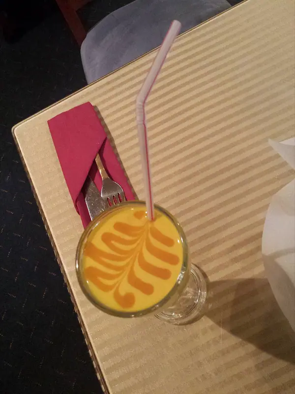 Overhead view of a yellow mango drink with an orange feather swirl and a striped straw, beside cutlery wrapped in a red napkin