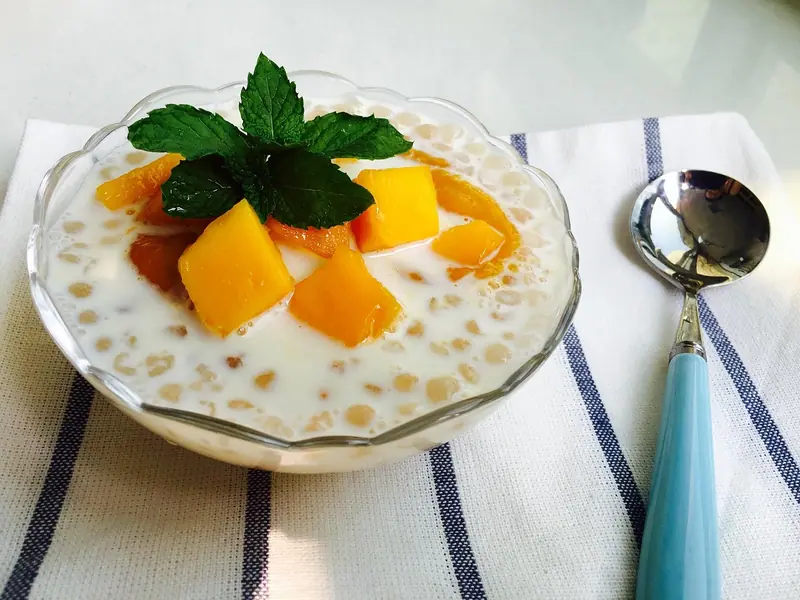 Glass bowl of creamy white milk with sago pearls, topped with mango cubes and a mint sprig, beside a blue-handled spoon on a striped cloth