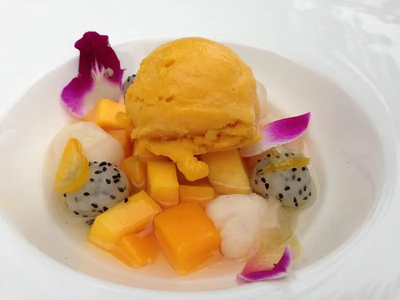 Eine Kugel orangefarbenes Mango-Sorbet in einer weißen Schale mit Mangowürfeln, Litschis, Drachenfruchtkugeln, Kumquatscheiben und lila Orchideenblüten.