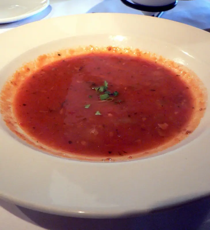 En bred hvid skål med tynd, rød tomatchowder med små stykker og krydderurter, toppet med lidt frisk persille i midten.