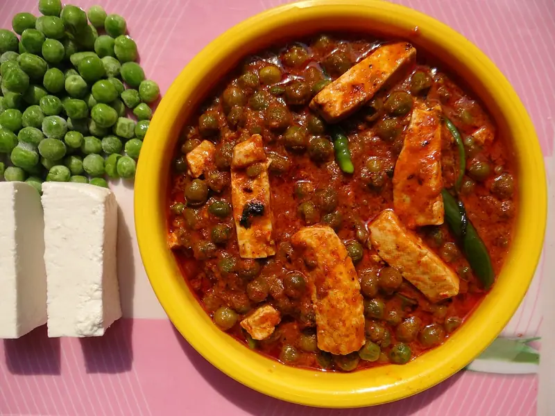 Yellow bowl of red-orange pea and paneer curry with slit green chillies, beside raw green peas and a block of paneer on a pink mat.