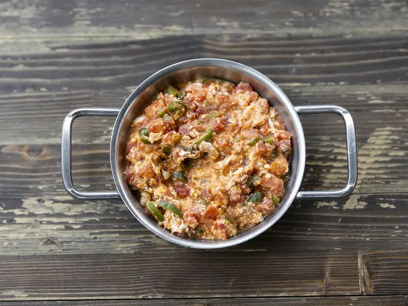 Scrambled eggs with chopped tomatoes and green pepper in a small two-handled stainless steel pan on a weathered wooden table.