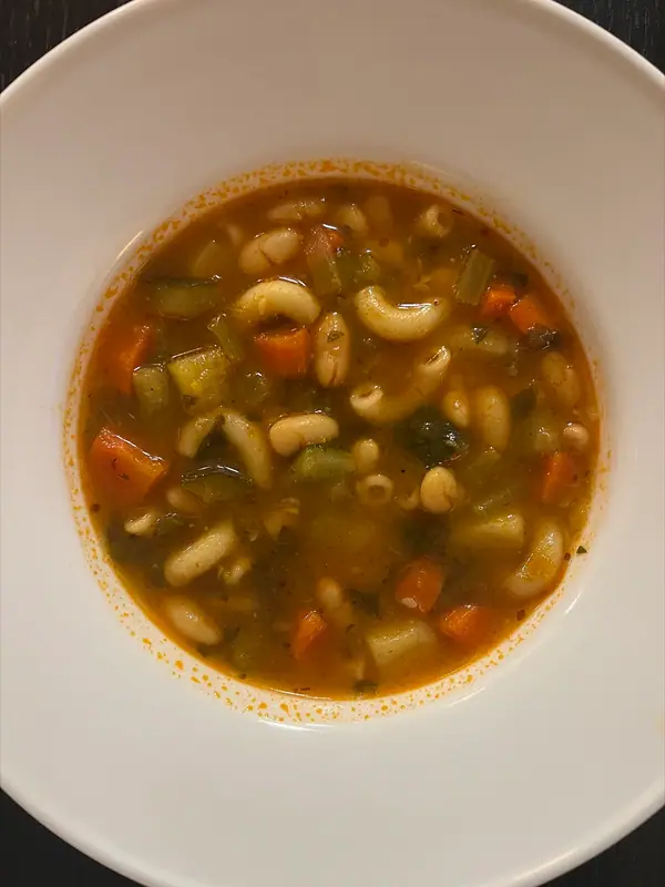 Sopa minestrone em uma tigela branca larga com macarrão cotovelo, feijão-branco, cenoura, abobrinha e folhas verdes em caldo de tomate, vista de cima.