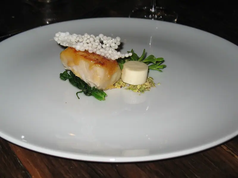 Seared white fish fillet topped with a puffed tapioca cracker, on wilted greens with a white cylinder and green crumble on a white plate