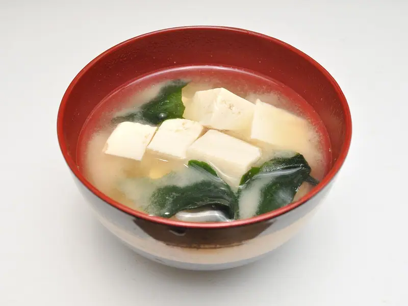 Bol roșu lăcuit cu supă miso tulbure, cuburi de tofu alb și alge wakame verde închis, pe o suprafață albă simplă.