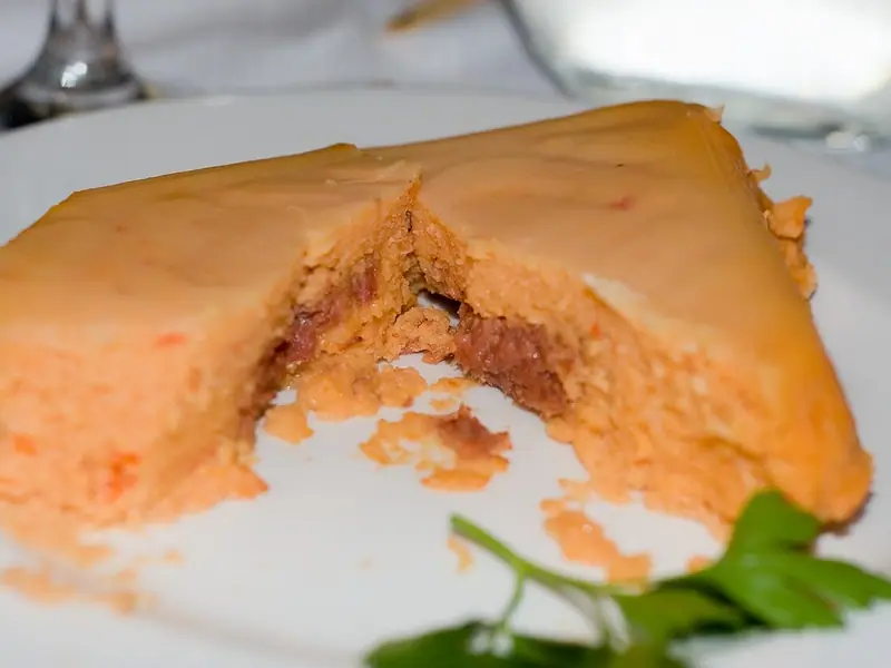 Trójkątny pomarańczowy pudding z fasoli przełamany na białym talerzu, odsłaniający ciemniejsze nadzienie, obok gałązka natki pietruszki.