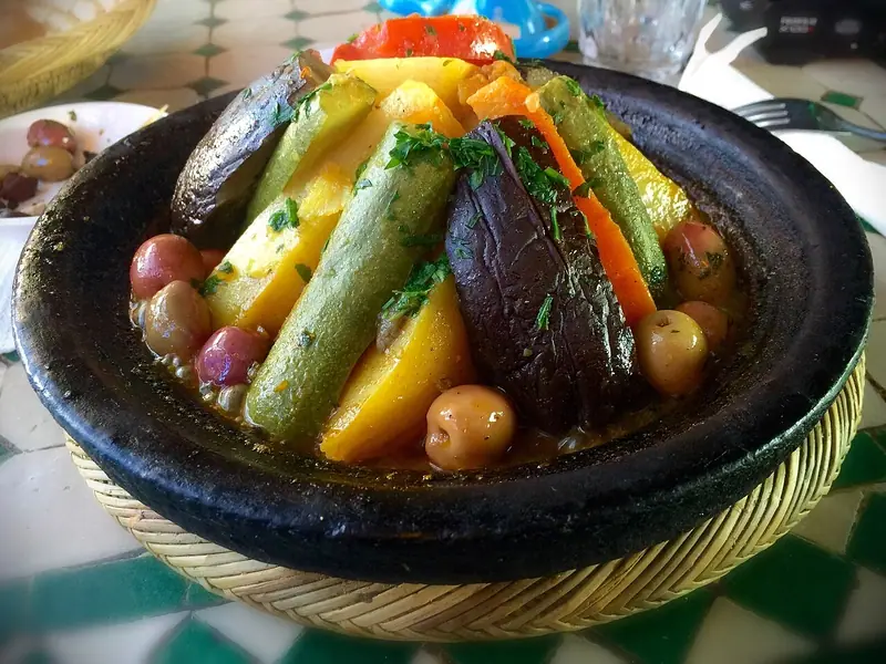 Grønnsakstagine i et svart leirfat med stående squash, aubergine, potet, gulrot og tomat, oliven og persille, på et flettet underlag